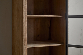 A close-up of the nkuku Nirvi Mango Wood & Iron Tall Cabinet - Black, featuring three shelves, stands empty by a glass door with a dark metal frame against a plain light-colored wall.