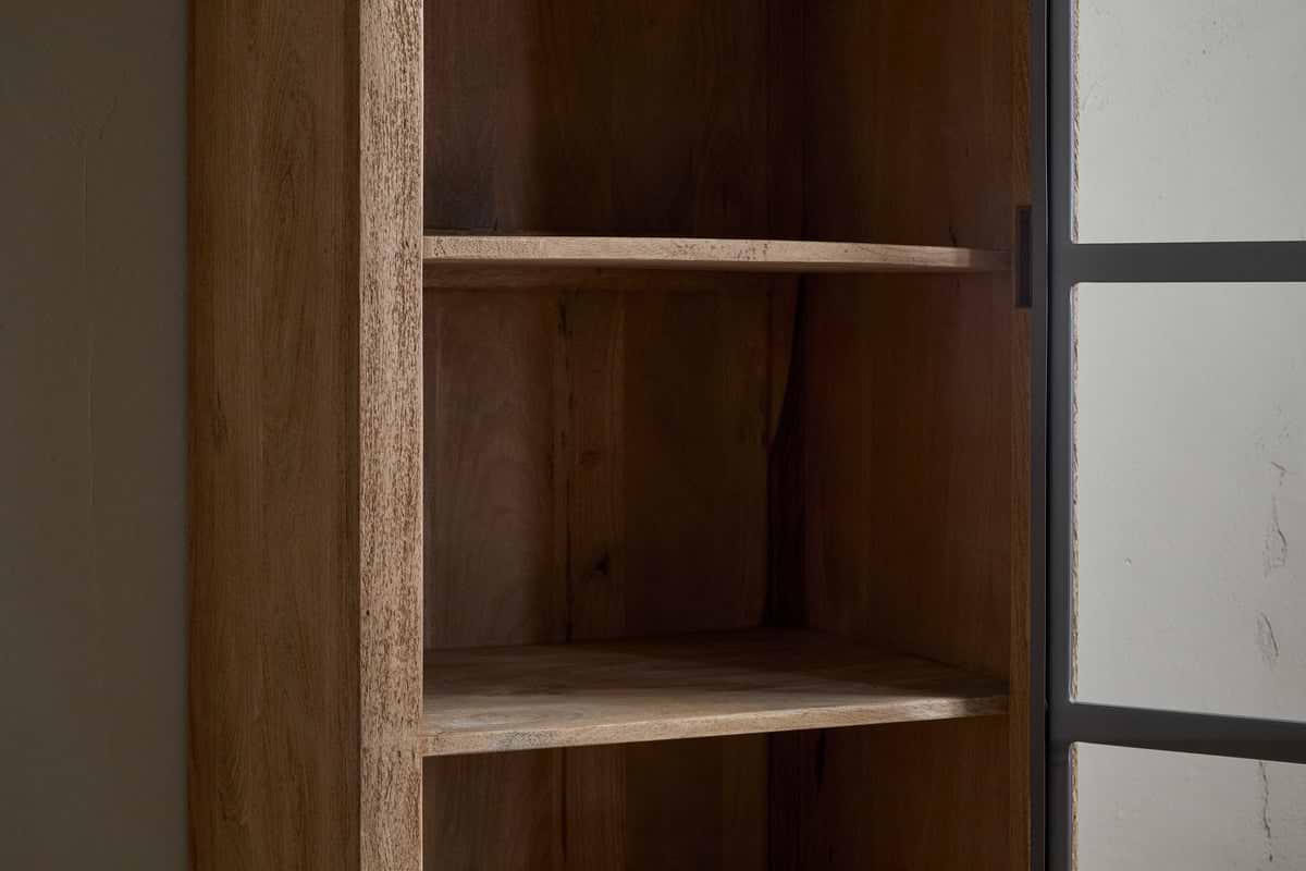 A close-up of the nkuku Nirvi Mango Wood & Iron Tall Cabinet - Black, featuring three shelves, stands empty by a glass door with a dark metal frame against a plain light-colored wall.