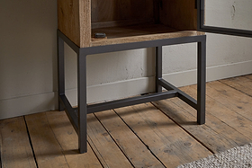 A close-up of the Nirvi Mango Wood & Iron Tall Cabinet - Black by nkuku, featuring a mango wood body and black iron frame, standing on a wooden floor beside a white wall and a beige textured rug.