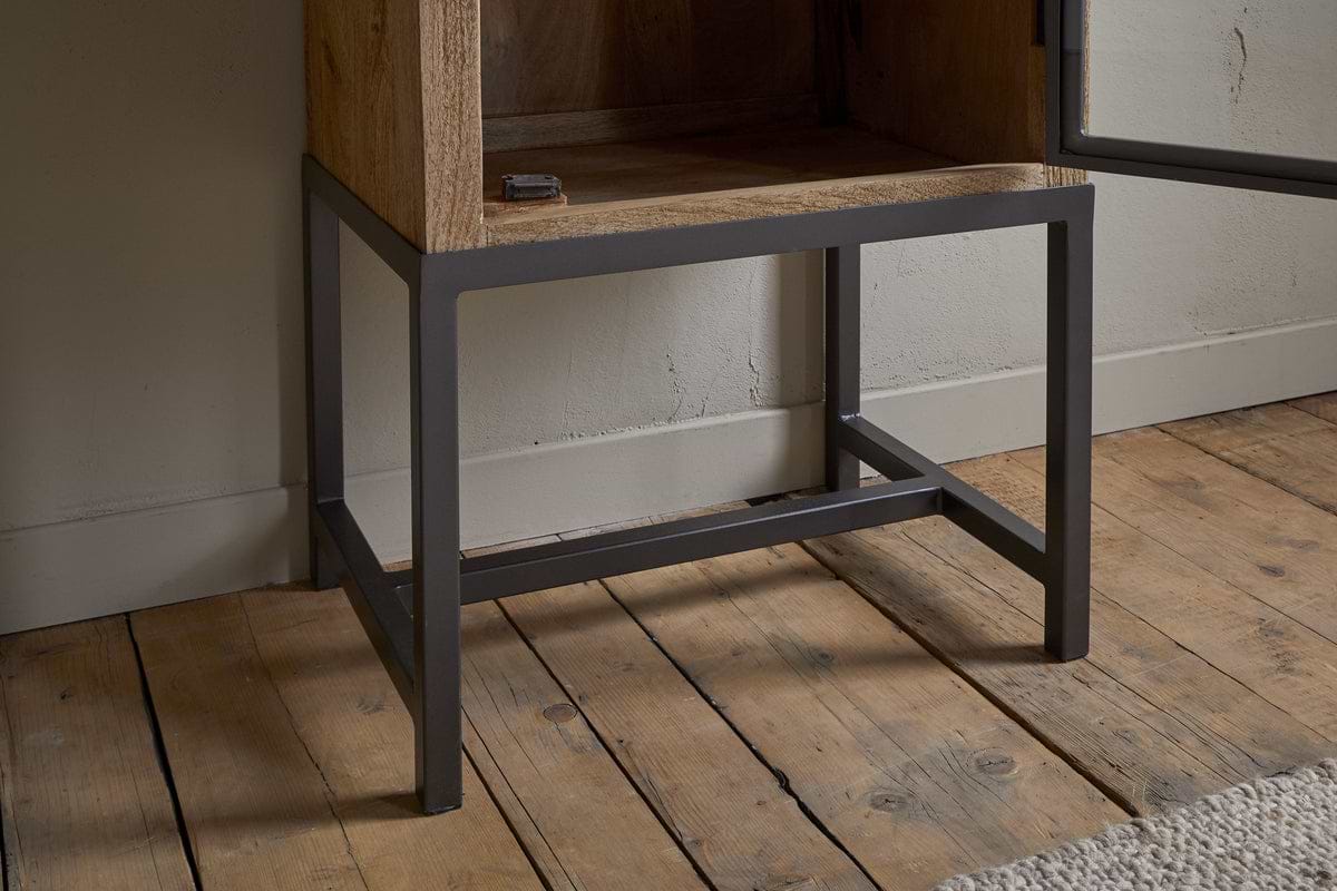 A close-up of the Nirvi Mango Wood & Iron Tall Cabinet - Black by nkuku, featuring a mango wood body and black iron frame, standing on a wooden floor beside a white wall and a beige textured rug.