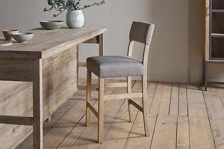 The Nadeeka Linen Upholstered Counter Chair by nkuku, in soft grey, sits beside a rustic wooden table on a light wood floor, topped with ceramic bowls and a vase with greenery—perfect for your kitchen island.