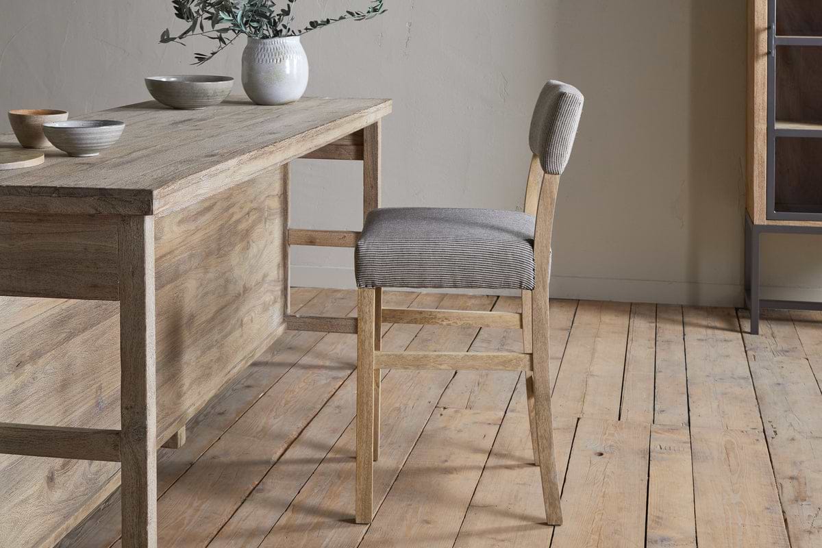 The nkuku Nadeeka Linen Upholstered Counter Chair in Soft Grey sits at a light wood dining table with ceramic bowls and a vase of greenery on top, all arranged on a wooden floor in a minimalist room.