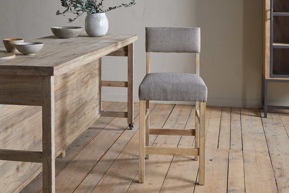 A wooden dining table with ceramic bowls and a white vase sits on a wooden floor next to the nkuku Nadeeka Linen Upholstered Counter Chair in Soft Grey, featuring linen upholstery and a supportive backrest.