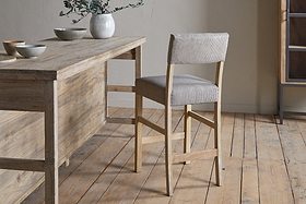 The nkuku Nadeeka Linen Upholstered Counter Chair in Soft Grey, featuring a cushioned backrest, stands by a rustic dining table on a light wood floor holding ceramic bowls and a vase with greenery.
