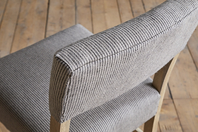 A close-up of the nkuku Nadeeka Linen Upholstered Counter Chair in Soft Grey, featuring striped linen fabric and a wooden frame, shown on a plank wooden floor.