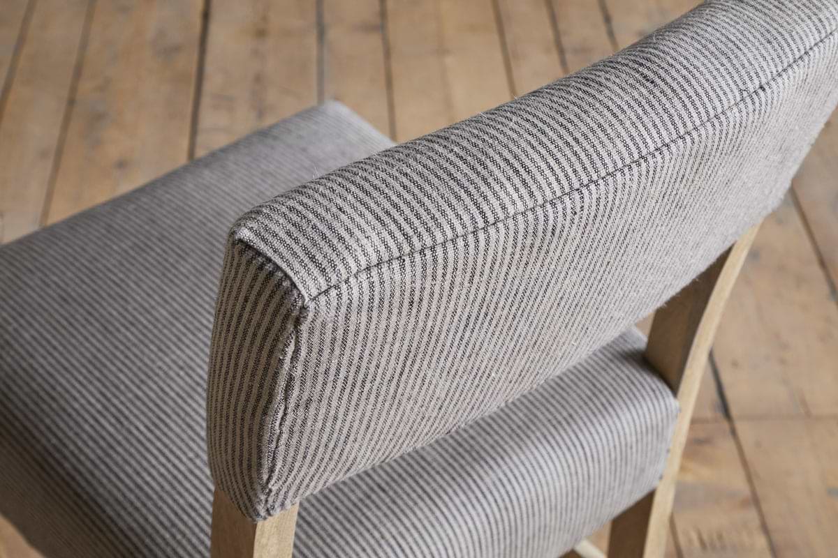 A close-up of the nkuku Nadeeka Linen Upholstered Counter Chair in Soft Grey, featuring striped linen fabric and a wooden frame, shown on a plank wooden floor.
