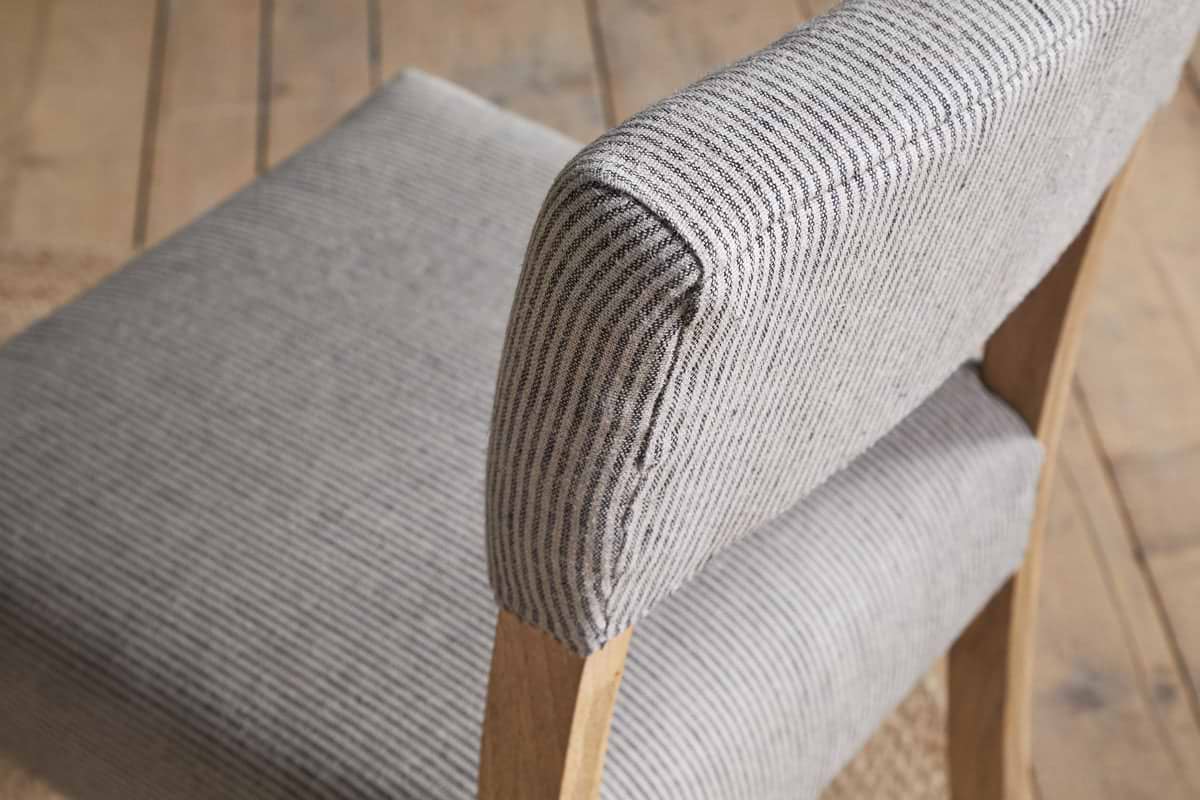 Close-up of the nkuku Nadeeka Linen Upholstered Dining Chair - Soft Grey, with striped linen fabric, cushioned seat and backrest, and light wood legs on a wooden floor.
