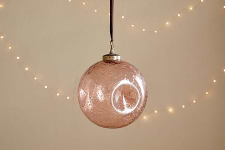The nkuku Nishan Pinched Giant Bauble - Copper is a large, round, recycled glass ornament with a metallic cap and brown ribbon, featuring a slightly speckled finish. Soft white fairy lights in the background add festive warmth.
