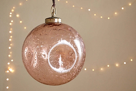 The nkuku Nishan Pinched Giant Bauble - Copper, crafted from recycled glass with beautiful bubbles inside, hangs elegantly from a ribbon. Warm white fairy lights blur in the background, creating a cozy, festive atmosphere as the bauble reflects light.