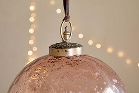 A close-up of nkuku’s Nishan Pinched Giant Bauble - Copper, made from textured recycled glass, hangs by a brown ribbon. Its metallic cap features a decorative loop, with warm white fairy lights softly glowing in the blurred background.