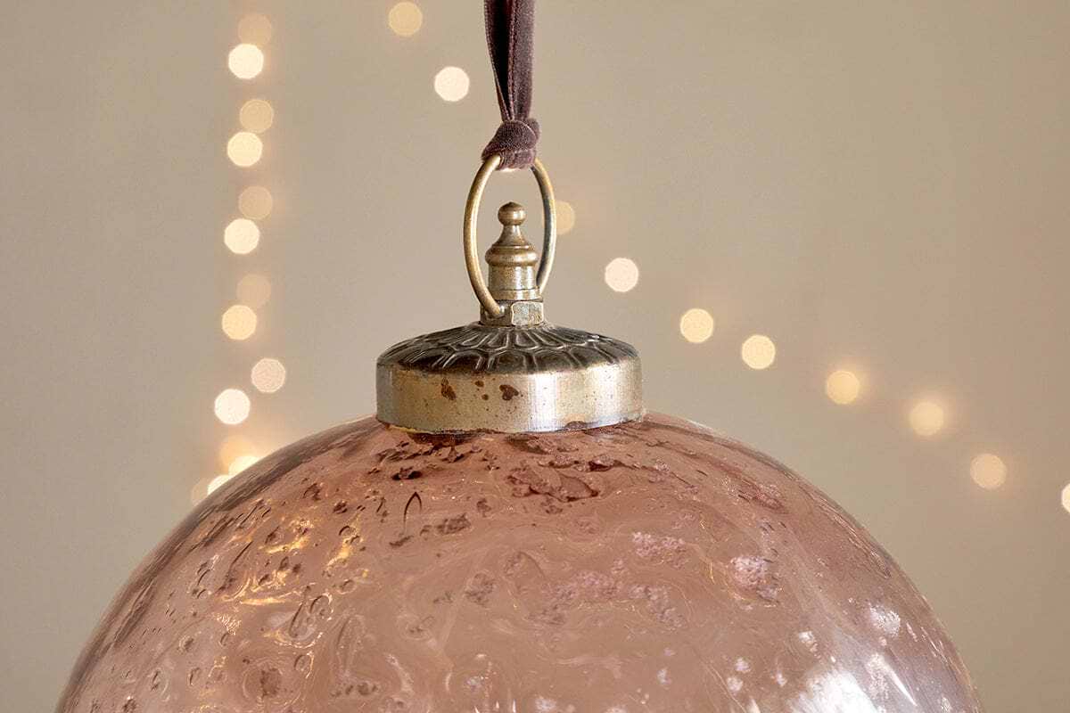 A close-up of nkuku’s Nishan Pinched Giant Bauble - Copper, made from textured recycled glass, hangs by a brown ribbon. Its metallic cap features a decorative loop, with warm white fairy lights softly glowing in the blurred background.