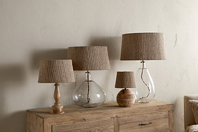 Four Nadee Banana Leaf Lampshades by nkuku, featuring natural textured shades atop glass or wooden bases, are arranged on a rustic wooden sideboard against a neutral wall, creating a cozy and organic look.