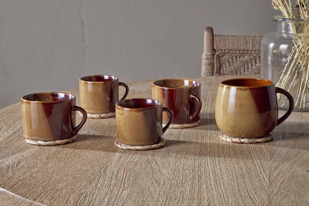 Five Naav Mustard Extra Large Mugs by nkuku rest on woven coasters atop a wooden table. A glass jar with dried plants and a wooden chair with a woven backrest are in the background, completing the scene.