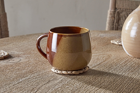 The Naav Mug - Mustard (Extra Large) by nkuku sits on a round woven coaster on a wooden table, with part of a vase and two woven placemats in the background.