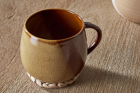 The Naav Mug - Mustard (Extra Large) by nkuku is a round ceramic mug with a brown and beige reactive glaze, paired with a woven coaster on a textured wooden surface.