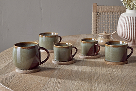 Two Naav Mugs - Blue (Small, Set of 2) by nkuku sit on wooden coasters atop a round table. In the background, a woven-back chair and vase of dried flowers enhance the cozy, rustic ambiance.