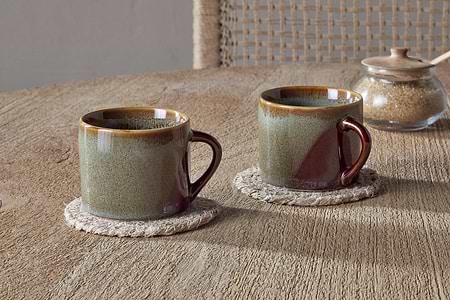 Two Naav Mugs - Blue - Small (Set of 2) by nkuku, featuring a unique two-tone reactive glaze, sit on woven coasters atop a wooden table. In the background are a glass jar of brown sugar, a small spoon, and a textured chair.