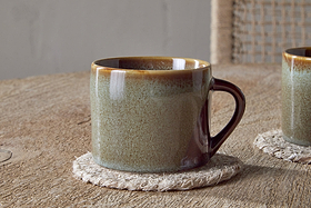A pair of Naav Mugs - Blue - Small (Set of 2) by nkuku rests on a rustic wooden table with woven coasters; one mug is partially visible, and a woven-back chair completes the charming tableau.