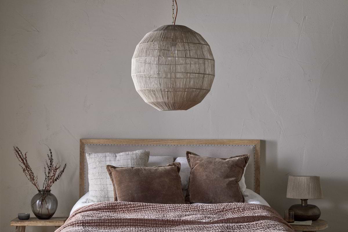 A cozy bedroom includes a wooden bed with white and brown bedding, two brown pillows, and the Nilaveli Woven Jute Sphere Pendant in Natural by nkuku hanging overhead. Wooden nightstands hold a small lamp and vase; the wall is light gray.