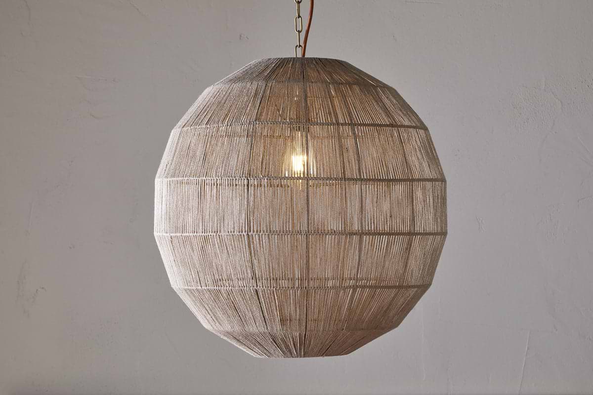 The nkuku Nilaveli Woven Jute Sphere Pendant in Natural hangs from the ceiling, its geometric, spherical jute shade showcasing a textured, see-through design that reveals the glowing bulb against a light-colored wall.
