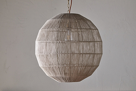 The Nilaveli Woven Jute Sphere Pendant by nkuku hangs from the ceiling, its natural jute threads tightly woven around a geometric frame. The textured, semi-transparent shade stands out against a plain, light-colored wall.