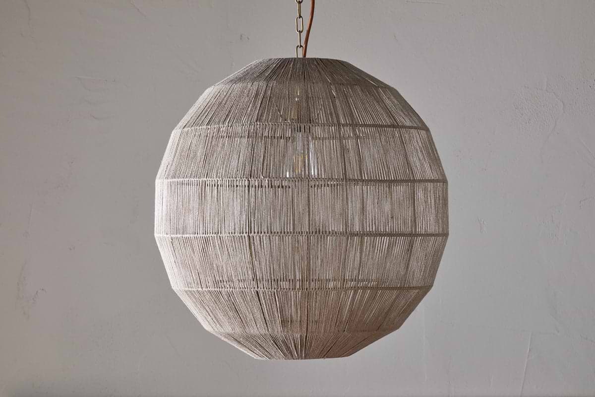 The Nilaveli Woven Jute Sphere Pendant by nkuku hangs from the ceiling, its natural jute threads tightly woven around a geometric frame. The textured, semi-transparent shade stands out against a plain, light-colored wall.