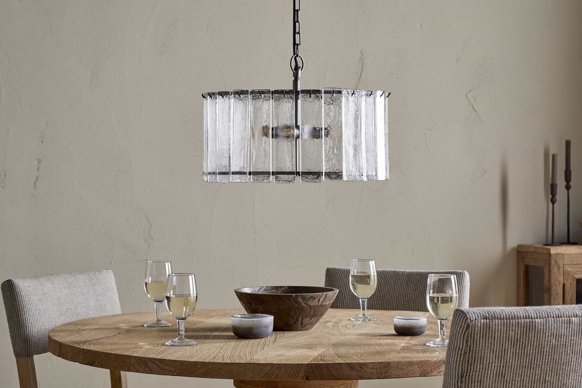 The Nauka Statement Pendant by nkuku, crafted from recycled glass with an aged bronze finish, elegantly hangs from the ceiling, making it a striking centerpiece above a table set with wine glasses.