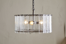 The Nauka Statement Pendant from nkuku features vertical recycled glass panels forming a round shade around three bulbs. Suspended by a black chain, this aged bronze and clear chandelier creates an elegant focal point on any light wall.