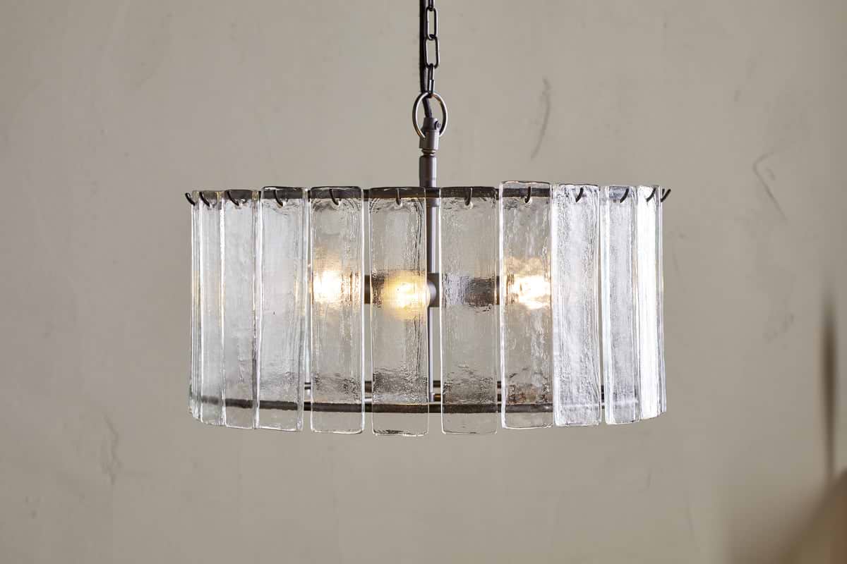 The Nauka Statement Pendant from nkuku features vertical recycled glass panels forming a round shade around three bulbs. Suspended by a black chain, this aged bronze and clear chandelier creates an elegant focal point on any light wall.