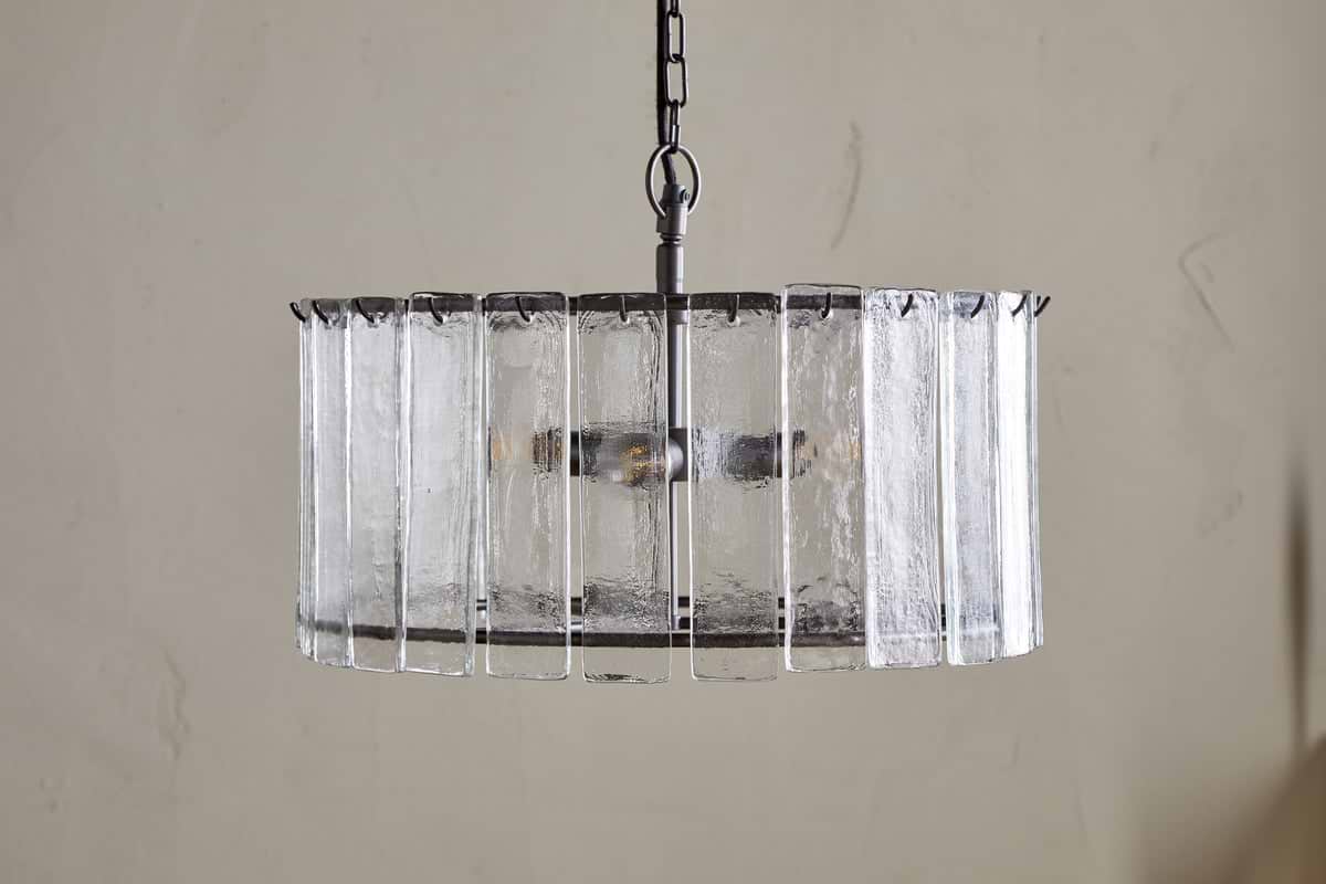 The nkuku Nauka Statement Pendant in Aged Bronze & Clear hangs from a chain, showcasing vertical textured recycled glass panels around its metal frame. Visible bulbs add elegant statement lighting against a light-colored wall.