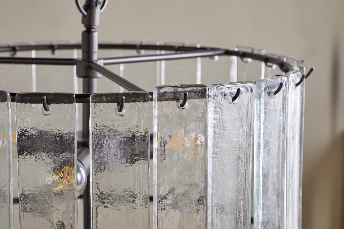 A close-up of the nkuku Nauka Statement Pendant in aged bronze and clear: vertically hanging textured recycled glass panels partly obscure glowing bulbs, creating a softly diffused light effect within a dark metal frame.