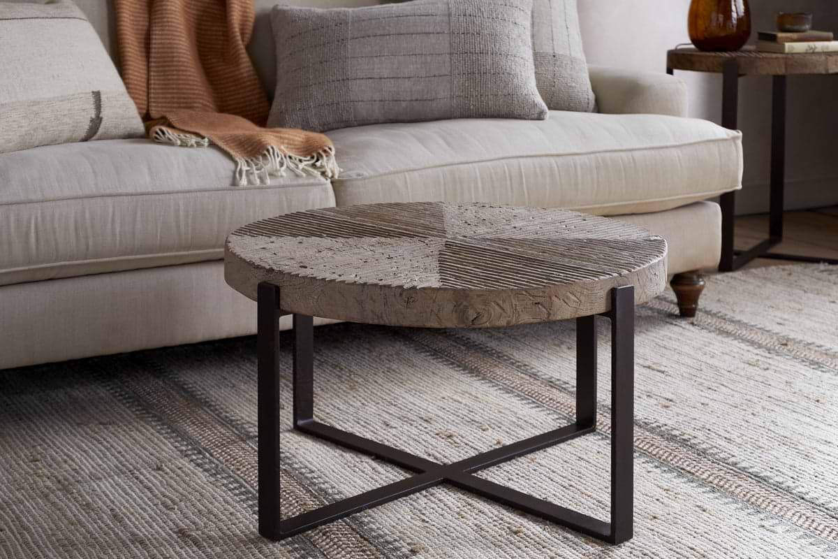The Chakala Reclaimed Wood Coffee Table by nkuku, featuring an iron base and reclaimed wood top, sits on a woven rug in front of a beige sofa with neutral and orange knit pillows. A side table and an amber glass vase appear in the background.
