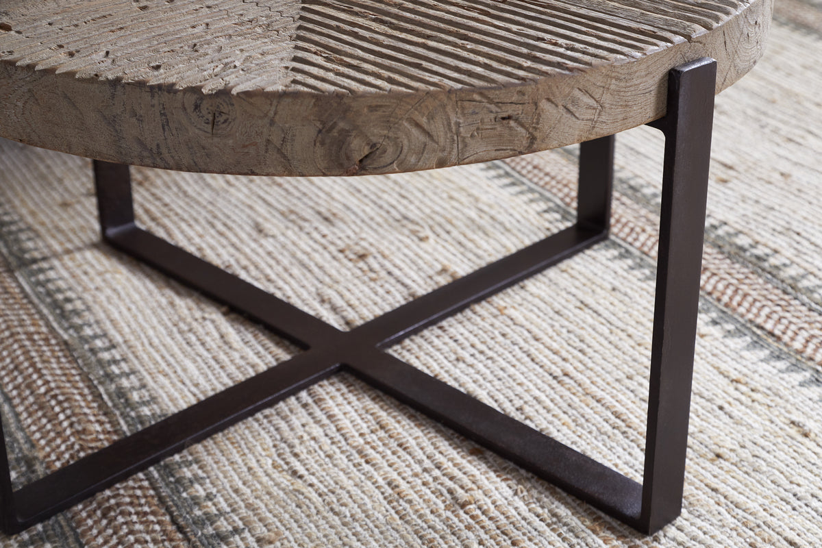 Close-up of the Chakala Reclaimed Wood Coffee Table by nkuku, featuring a textured reclaimed wood top and black iron X-shaped base, set atop a woven beige rug with brown and green striped patterns.