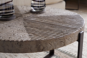 A close-up of the nkuku Chakala Reclaimed Wood Coffee Table, showcasing its geometric carved top in reclaimed wood and iron legs. Two glass vases with dark stripes sit on it, all resting above a neutral textured rug.