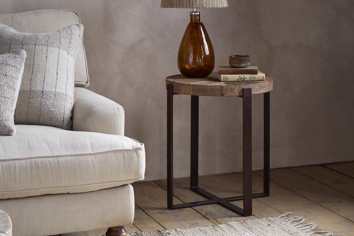 A cozy beige armchair with patterned cushions sits beside the nkuku Chakala Reclaimed Wood Side Table, crafted from reclaimed wood and iron. The table holds stacked books, a brown glass lamp with a woven shade, and a small bowl atop a woven rug.