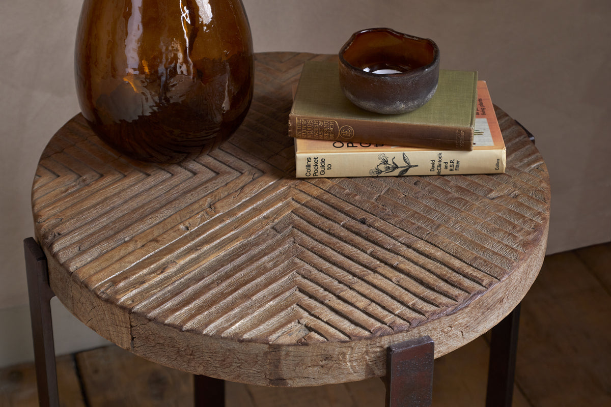 The nkuku Chakala Reclaimed Wood Side Table features a herringbone pattern and holds a brown glass vase, small ceramic bowl, and two stacked hardcover books. It's set indoors on a wooden floor with a neutral wall background.