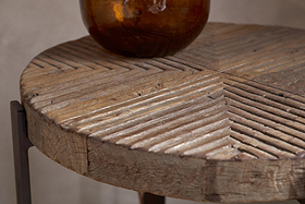 A close-up of the Chakala Reclaimed Wood Side Table by nkuku showcases its carved diagonal grooves and weathered wood. Iron legs and an amber glass vase highlight the sustainable elegance of this reclaimed wood piece.