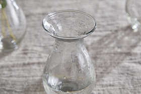 The nkuku Bapti Bud Vases - Clear (Set of 3) feature wide, flared rims and narrow necks. One vase with visible water droplets stands on a light gray textured surface, while two blurred vases are in the background.