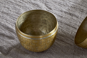 The Mutti Hammered Opening Egg by nkuku, crafted from antique brass with intricate engravings and a softly aged finish, rests on textured gray fabric. A glimpse of a second similar piece appears at the edge, making it a unique seasonal keepsake.