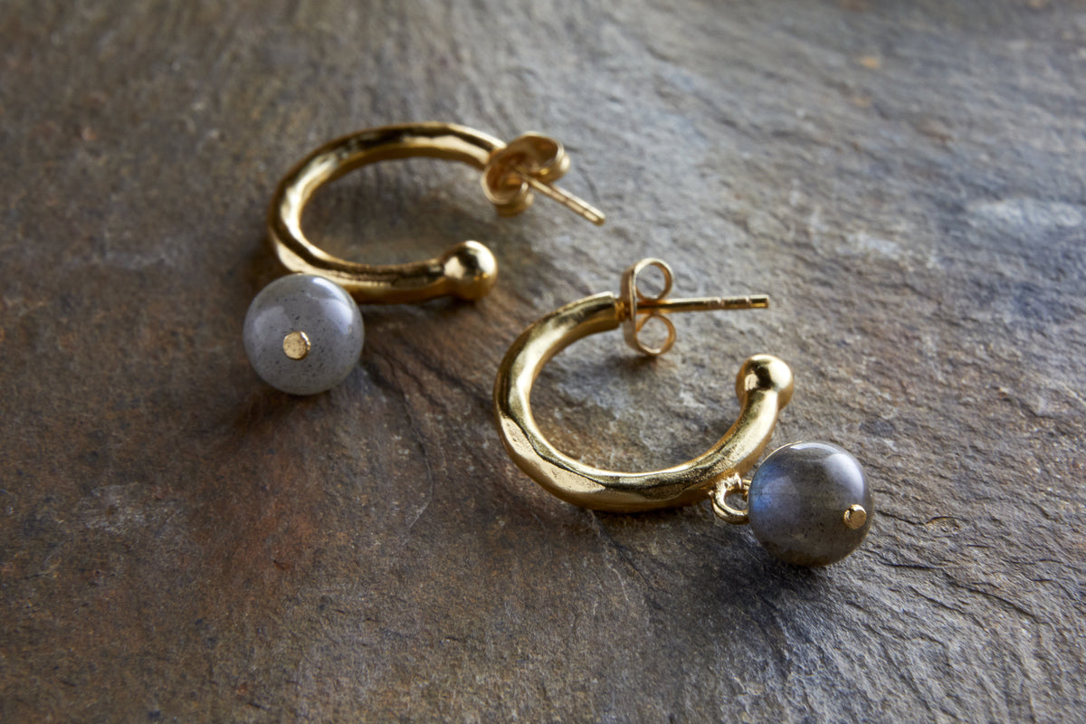 The Marai Labradorite Earrings by nkuku feature gold hoops with organic curves and a round, grayish labradorite bead drop, creating an elegant silhouette—an ideal jewelry gift.