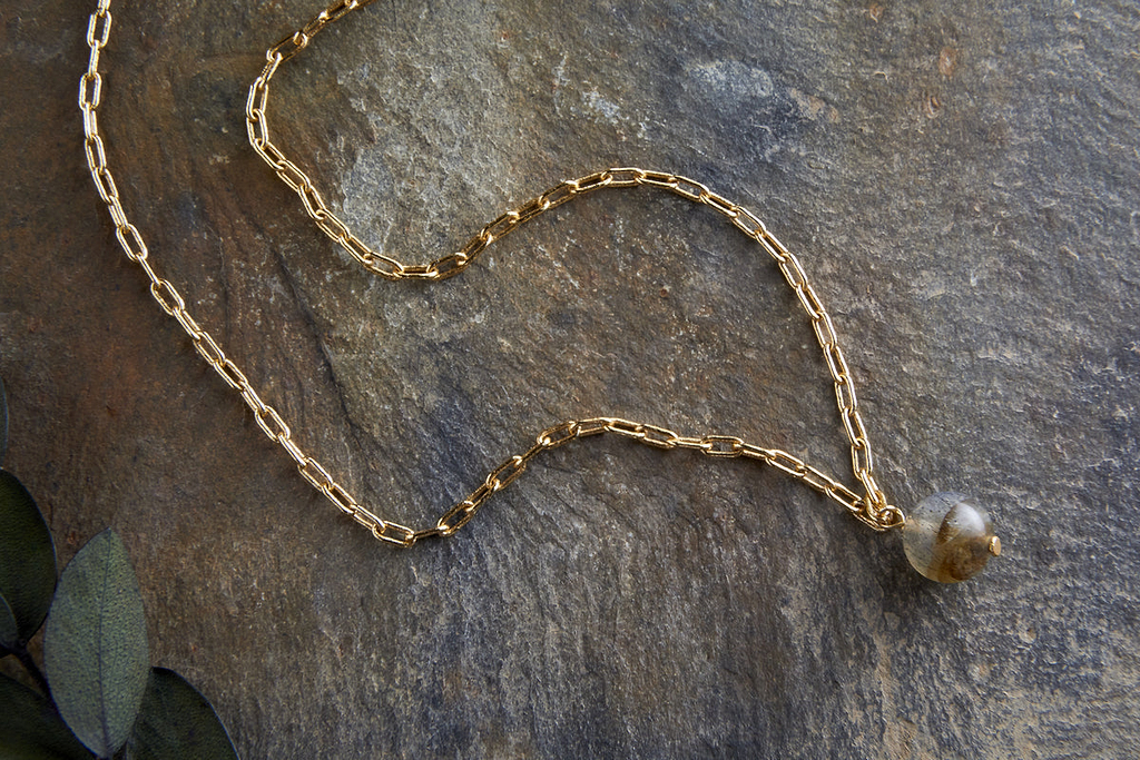 The Marai Labradorite Necklace - Labradorite & Gold by nkuku features an elongated gold chain with a grayish labradorite pendant, displayed on textured stone with green leaves, suggesting it as an elegant jewelry gift.
