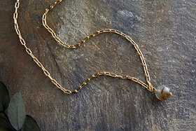 The Marai Labradorite Necklace - Labradorite & Gold by nkuku features an elongated gold chain with a grayish labradorite pendant, displayed on textured stone with green leaves, suggesting it as an elegant jewelry gift.