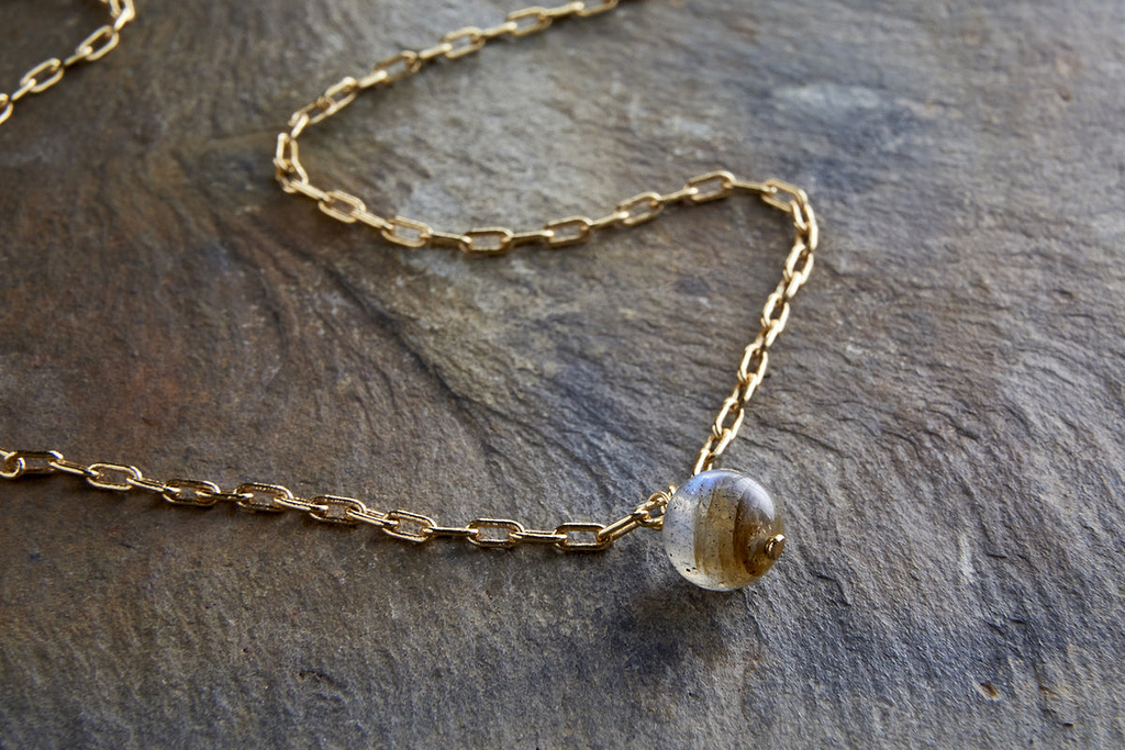 The Marai Labradorite Necklace by nkuku features a gold-plated pendant and a round labradorite bead with subtle gold and brown streaks, creating a warm look. Displayed on textured stone, it’s an elegant jewellery gift.