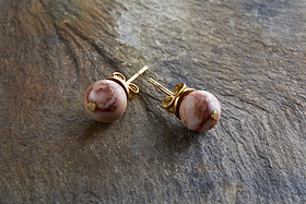 The Cerita Rhodochrosite Stud Earrings by nkuku feature round, marbled pink and white rhodochrosite stones in gold-plated settings, showcased on a textured brown stone surface for an elegant look.