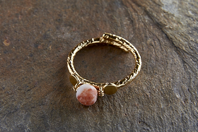 The Cerita Rhodochrosite Ring by nkuku features a twisted, gold-plated band displayed on stone, showcasing a round pinkish-orange rhodochrosite with white marbling, held by wire and flanked by two small gold discs.