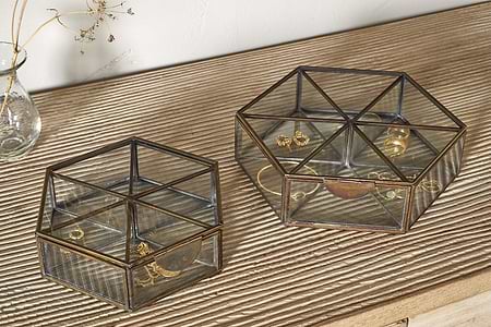 Two nkuku Sitara Jewellery Boxes in antique brass sit on a ribbed wooden surface. The smaller holds a gold bracelet and ring, the larger has earrings, rings, and a necklace. A glass vase with dried flowers is nearby.