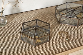 On a wooden surface, two nkuku Sitara Jewellery Boxes in antique brass—one square, one hexagonal—hold gold jewelry. Beside them lies a gold bracelet with a green charm. A glass vase with dried flowers is in the upper left corner.