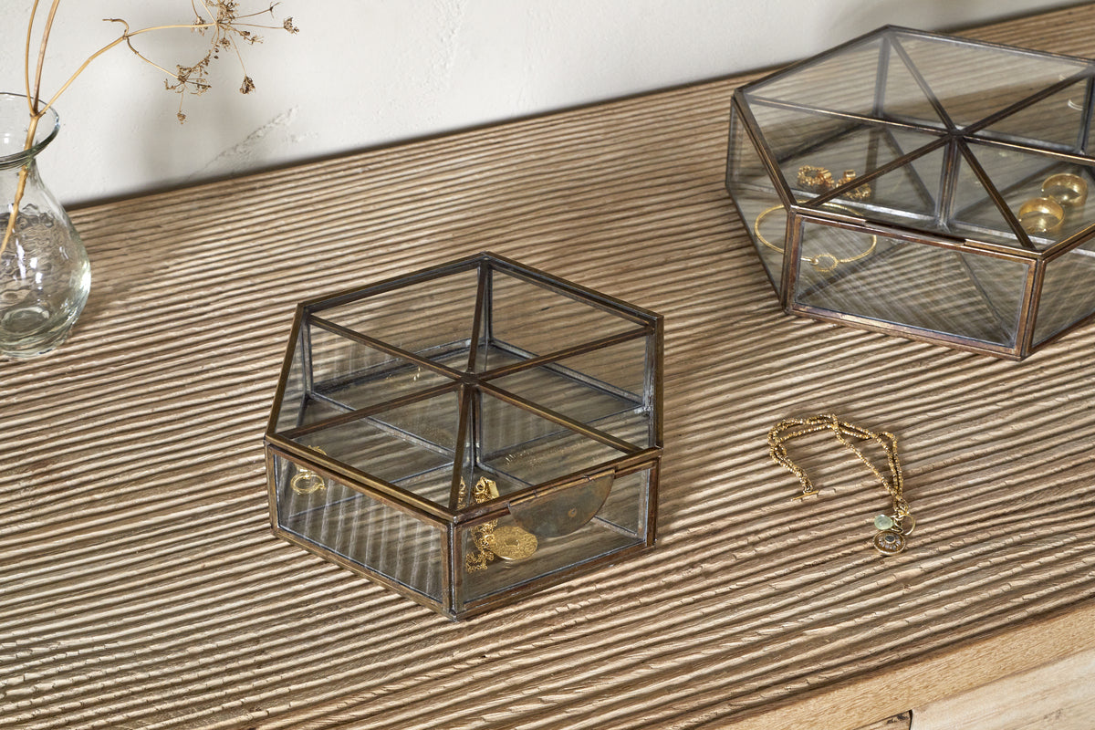 On a wooden surface, two nkuku Sitara Jewellery Boxes in antique brass—one square, one hexagonal—hold gold jewelry. Beside them lies a gold bracelet with a green charm. A glass vase with dried flowers is in the upper left corner.