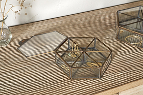 The Sitara Jewellery Box - Antique Brass by nkuku sits open on a ribbed wooden surface, displaying assorted gold rings and bracelets beside a small vase of dried flowers and another closed glass box.