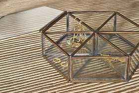 The Sitara Jewellery Box - Antique Brass by nkuku sits open on a ribbed beige surface, displaying gold necklaces, rings, and a green gemstone bracelet neatly inside, their reflections shimmering on the glass sides and base.